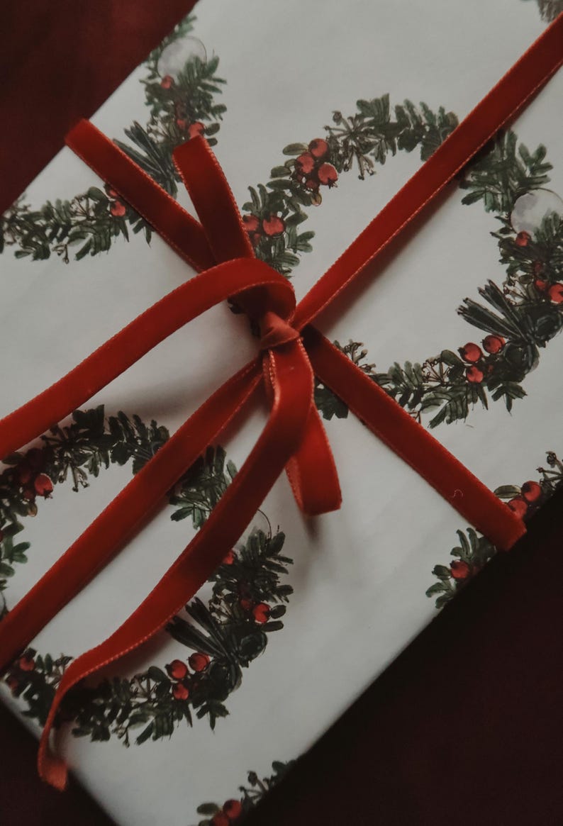 May include: A white gift box with a red velvet ribbon tied in a bow. The box is wrapped in paper with a repeating pattern of green wreaths with red berries.