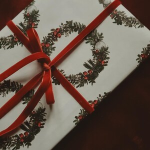 May include: A white gift box with a red ribbon tied in a bow. The box is wrapped in paper with a repeating pattern of green wreaths with red berries.