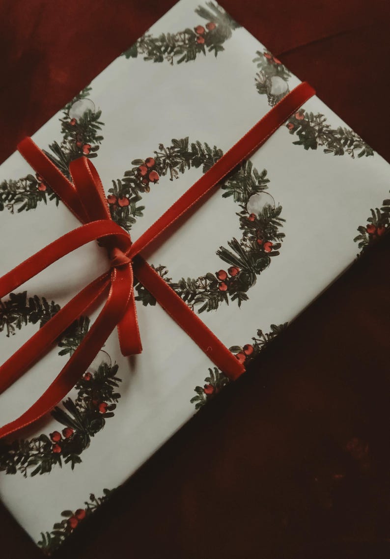 May include: A white gift box with a red ribbon tied in a bow. The box is wrapped in paper with a repeating pattern of green wreaths with red berries.