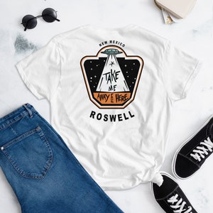 Pode incluir: Camiseta branca com um gráfico preto e laranja de um OVNI com o texto "Take Me Away From Here" e "Roswell, Novo México".