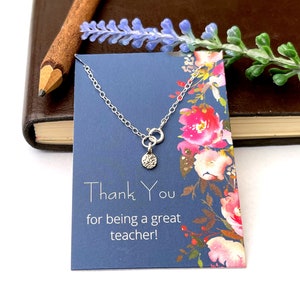 Könnte beinhalten: Eine silberfarbene Kettenhalskette mit einem kleinen, kreisförmigen, strukturierten Anhänger auf einer blauen Präsentationskarte. Die Karte zeigt ein Blumenmuster und den Text: Thank You for being a great teacher! Die Kette hat einen Federringverschluss.
