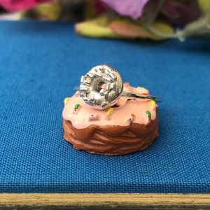 May include: A silver ring featuring a donut design. The ring is displayed on a pink iced donut with colourful sprinkles.