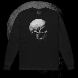 May include: Black long-sleeve shirt featuring a detailed, grayscale image of a human skull. The skull is centered on the shirt, with a dark background. The design has a distressed, textured appearance.