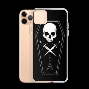 Puede incluir: Carcasa de tel&eacute;fono negra con un dise&ntilde;o blanco de calavera y huesos cruzados dentro de un ata&uacute;d. El ata&uacute;d tiene un tri&aacute;ngulo y un s&iacute;mbolo de cruz.
