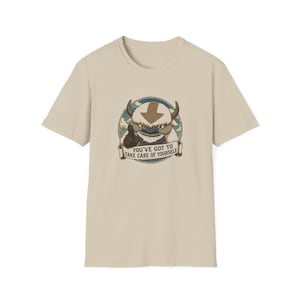 May include: A beige short-sleeve t-shirt featuring a circular graphic of a furry creature with horns and a brown arrow on its forehead. Below the creature, a banner contains the text: YOU'VE GOT TO TAKE CARE OF YOURSELF. The design is centered.