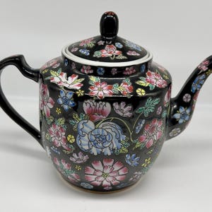May include: A black ceramic teapot featuring a vibrant floral pattern with pink, blue, and yellow flowers and green leaves. The teapot includes a matching lid with a black knob, a curved handle, and a spout, all decorated with the same floral motif.