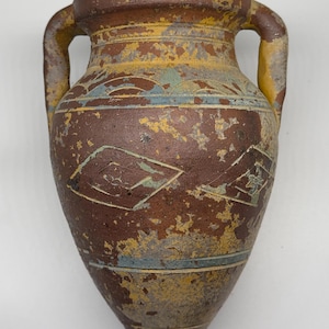 May include: A terracotta-colored amphora with two side handles, featuring a weathered finish with patches of yellow, blue, and white paint. The vessel displays geometric patterns, including diamond shapes with floral motifs and horizontal bands.