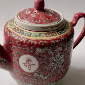 May include: A ceramic teapot featuring a vibrant red base colour with intricate white floral patterns and swirling scrollwork. The teapot includes a matching lid, a curved spout, and a handle. A white circular medallion displays a red Chinese character.