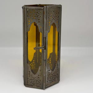 May include: A hexagonal lantern featuring a weathered metallic frame with intricate cut-out patterns at the top and bottom. The structure holds amber-colored glass panels and includes a simple metal latch mechanism on one of the vertical edges.