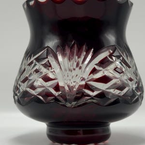 May include: A deep red glass vase featuring a scalloped rim and intricate clear cut-glass patterns. The design includes diamond-shaped facets and a central fan-like motif that reveals the clear glass beneath the dark red surface of the vessel.