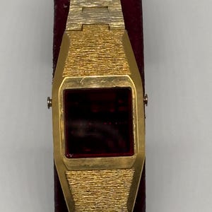 May include: A gold-tone digital wristwatch featuring a textured metal band and a rectangular case with a dark red display screen. The watch has a metallic finish with a distinct cross-hatch pattern on the links and the bezel surrounding the screen.