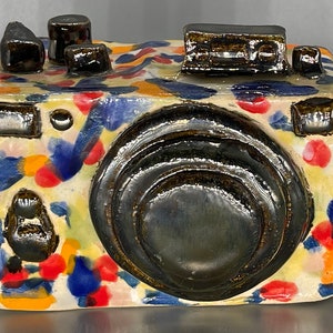May include: A ceramic camera with a colorful abstract pattern. The camera is painted in shades of blue, red, yellow, and white. The lens is black and has a glossy finish.