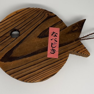 Può includere: Un sottopentola in legno a forma di pesce con venature naturali color marrone, un foro circolare vicino alla testa e un occhiello in corda marrone attaccato alla coda. Un'etichetta rettangolare rosa con testo giapponese è fissata al centro.