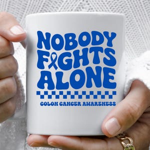 May include: White ceramic mug with the words "NOBODY FIGHTS ALONE" in blue, along with a blue ribbon and the text "COLON CANCER AWARENESS". The mug is held by a person.