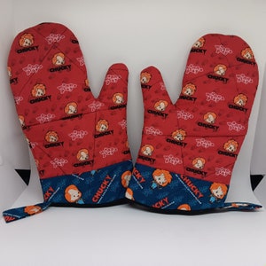 May include: A pair of red oven mitts with a blue trim. The mitts feature a repeating pattern of a cartoon character with the word "Chucky" and the phrase "Wanna Play".