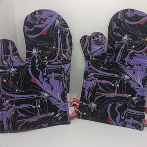 May include: A pair of oven mitts with a black background and a purple and red character design. The mitts are made of fabric and have a loop for hanging. The design features a stylized illustration of a character.