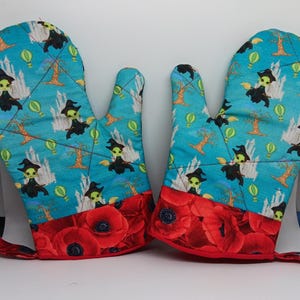 May include: A pair of blue and red oven mitts with a whimsical pattern of green witches flying on broomsticks and hot air balloons. The mitts have a poppy flower design on the bottom.