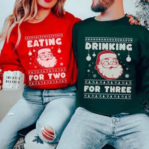 Op de afbeelding: Twee kersttruien, één rode en één groene. De rode trui zegt "EATING FOR TWO" met een kerstman-afbeelding. De groene trui zegt "DRINKING FOR THREE" met een kerstman-afbeelding. Beide truien hebben een feestelijk ontwerp.