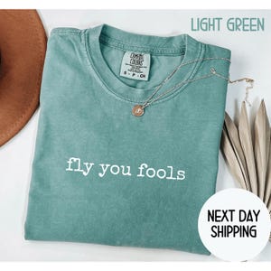 May include: Light green Comfort Colors t-shirt with the phrase "fly you fools" printed in white. A silver necklace with a pendant rests on the shirt. The shirt is a size small.