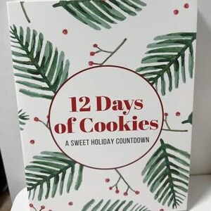 May include: A white box with a festive design featuring green fern-like leaves and red berries. The center has a circular design with the text "12 Days of Cookies" in red, with "A Sweet Holiday Countdown" below.