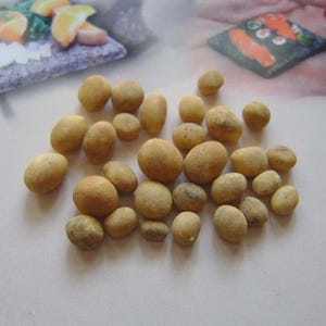 May include: A collection of small, light brown, oval-shaped objects, possibly decorative stones or food items, are scattered across a white surface. The objects vary slightly in size and shape, with a textured appearance. The background includes blurred images of food.