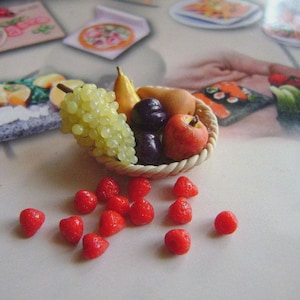Op de afbeelding: Een miniatuur gevlochten mandje gevuld met verschillende soorten fruit, waaronder groene druiven, een banaan, pruimen en een rode appel. Verspreid rond de mand liggen verschillende kleine rode aardbeien. De vruchten zijn op een lichtgekleurd oppervlak geplaatst.