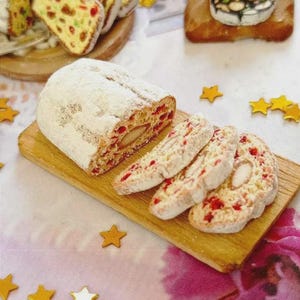 Puede incluir: Un pastel stollen festivo espolvoreado con azúcar glas, cortado para revelar el relleno de fruta, se presenta en una tabla de madera. También se ven rebanadas de stollen y otros productos horneados, junto con decoraciones en forma de estrella.