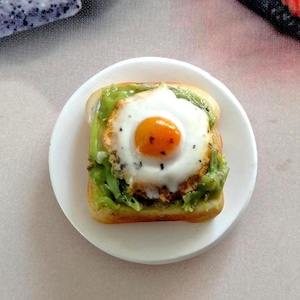 May include: A miniature breakfast of toast with avocado and a fried egg, presented on a small white plate. The egg yolk is a bright orange, and the avocado spread is a vibrant green. The toast is golden brown.