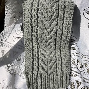 May include: A gray knitted scarf with a textured pattern. The scarf features a central chevron design and vertical ribbed sections. The soft, warm scarf is made of a light gray yarn and is suitable for cold weather.