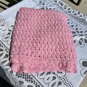 May include: A handmade, light pink crochet blanket. The blanket features a textured stitch pattern and a scalloped edge. The soft pink color and intricate design suggest a cozy, handcrafted item, perfect for a baby or as a decorative throw.
