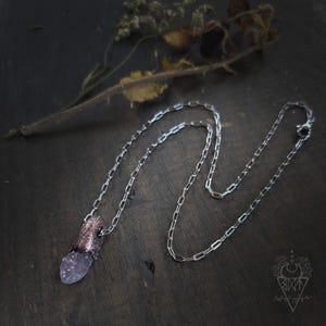 May include: A silver-toned chain necklace featuring a pendant with a textured, dark metallic cap and a translucent, pale purple crystal point. The chain consists of elongated rectangular links and includes a small lobster claw clasp closure.