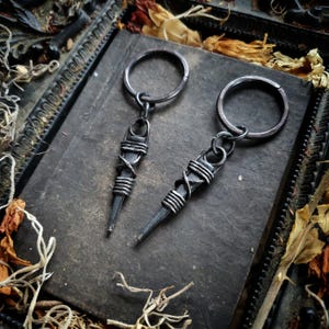 May include: Two silver keychains with a gothic aesthetic. Each keychain features a pointed, nail-like pendant wrapped with wire. The keyrings are circular and attached to the pendants with a small loop. The background is a dark, textured surface.
