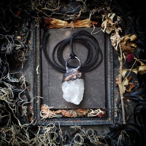 May include: A pendant necklace with a raw, clear quartz crystal. The crystal is set in a copper and silver-toned metal and suspended from a black cord. The necklace is presented on a dark, textured surface within a decorative frame, evoking a gothic aesthetic.