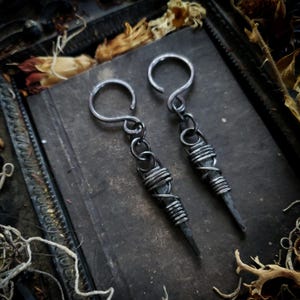 May include: A pair of silver earrings. Each earring has a long, pointed design wrapped with wire. The earrings have a hook closure and are displayed on a dark, textured surface.