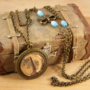 Peut inclure: Un collier en bronze avec un pendentif rond représentant une image brune et blanche. Le collier est posé sur une valise en cuir marron de style vintage avec des perles bleues et blanches.