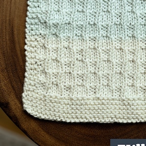May include: A close-up of a knitted dishcloth with a textured pattern in shades of blue, white, and cream. The dishcloth is made with a simple stitch and has a finished edge. The text "Full Video Tutorial" is visible at the bottom of the image.