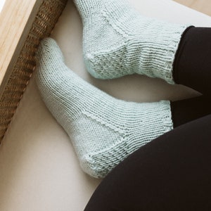 May include: A pair of pale blue knitted socks. The socks are worn on a person's feet, with the legs in black leggings. The socks have a textured pattern and are resting on a white surface. The background includes a wooden floor and a light-coloured chair.