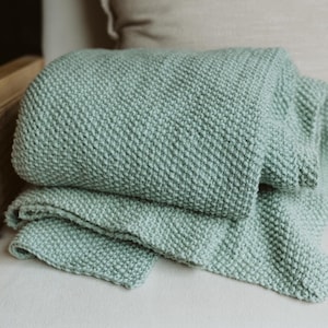 May include: A light green knitted throw blanket folded on a light beige chair with a light beige cushion.