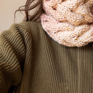 May include: A woman wearing a brown knitted jumper with a pink and beige knitted cowl neck scarf.
