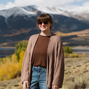 Puede incluir: Una mujer con un cárdigan marrón, una camiseta marrón y unos vaqueros azules se encuentra frente a una cordillera con un lago al fondo. Las montañas están cubiertas de nieve y los árboles están cambiando de color.