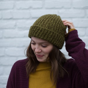 May include: A woman wearing a dark purple cardigan over a mustard yellow sweater and a green knitted beanie.