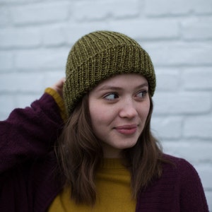 May include: A woman wearing a green knitted beanie, a mustard yellow sweater, and a burgundy cardigan. She is standing in front of a white brick wall.