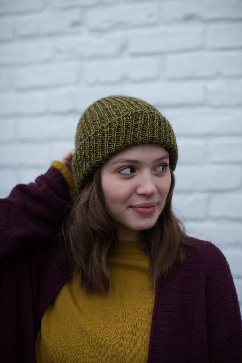 May include: A woman wearing a green knitted beanie, a mustard yellow sweater, and a burgundy cardigan. She is standing in front of a white brick wall.