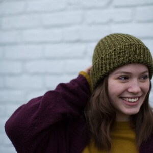May include: A woman wearing a green knit beanie, a burgundy cardigan, and a mustard yellow sweater. She is smiling and looking to the right.