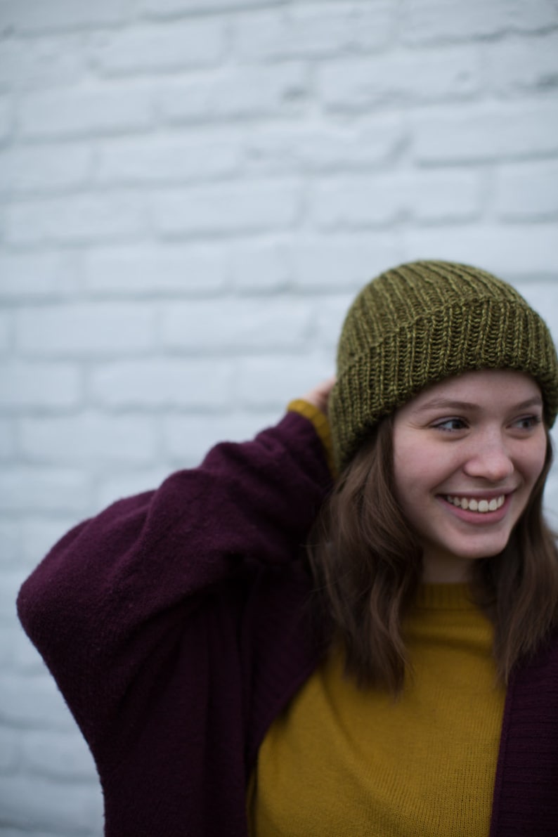 May include: A woman wearing a green knit beanie, a burgundy cardigan, and a mustard yellow sweater. She is smiling and looking to the right.