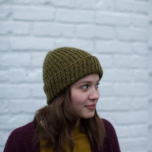 May include: A woman wearing a dark purple cardigan over a mustard yellow shirt and a green knitted beanie.
