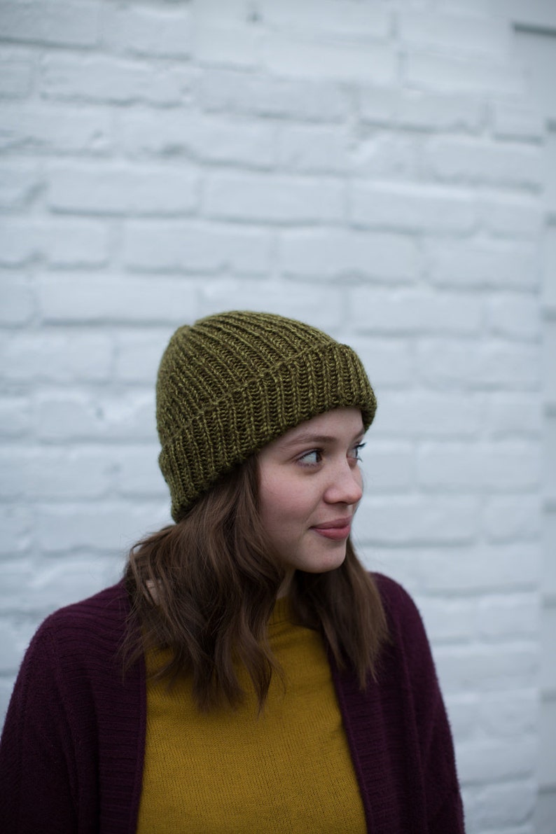 May include: A woman wearing a dark purple cardigan over a mustard yellow shirt and a green knitted beanie.