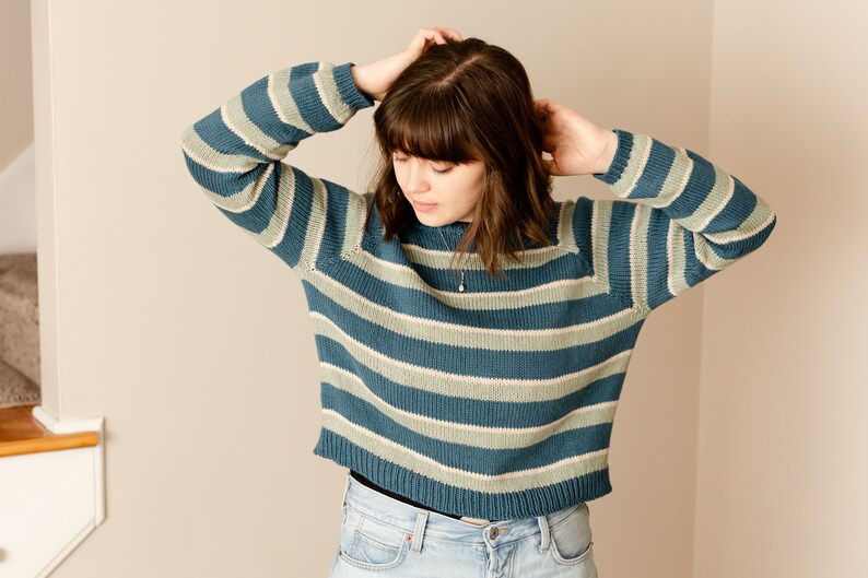 May include: A woman wearing a blue and green striped jumper with a delicate silver necklace. The jumper has a relaxed fit and a slightly cropped length.