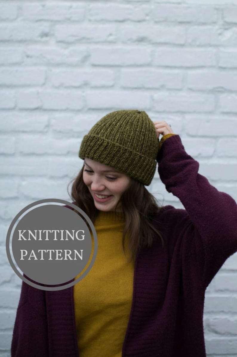May include: A woman wearing a dark purple cardigan over a mustard yellow turtleneck sweater and a green knitted beanie. The text "Knitting Pattern" is displayed in a circle.