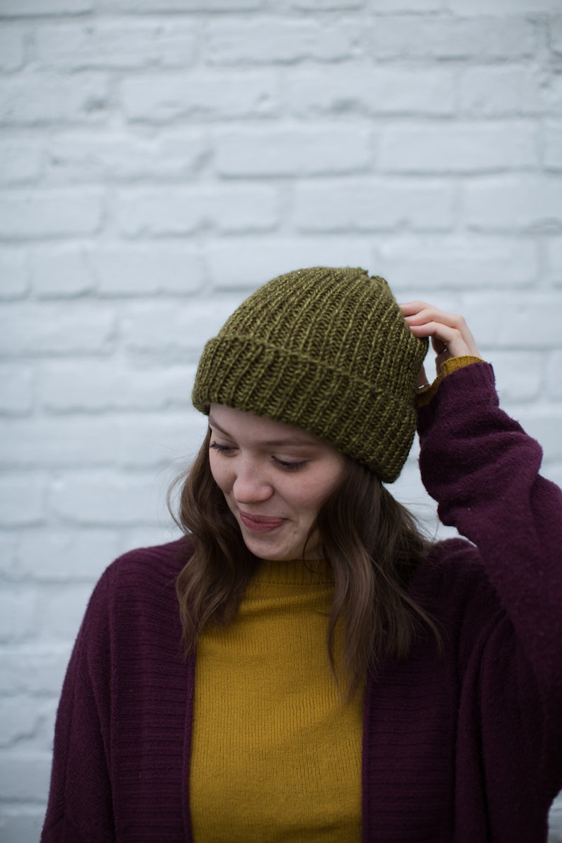May include: A woman wearing a dark purple cardigan over a mustard yellow sweater and a green knitted beanie.